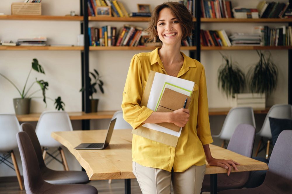 jeune femme avec un t shirt jaune tenant des dossiers et souriant, appuyée contre un bureau